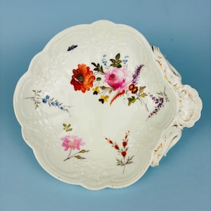 May include: A white porcelain shell-shaped dish featuring embossed scrollwork edges with gold trim. The interior surface is decorated with colourful hand-painted floral arrangements including red poppies, pink roses, and small blue and purple wildflowers.