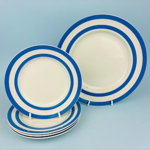 Puede incluir: Un juego de platos de cerámica de color crema con un llamativo patrón de bandas circulares de color azul oscuro. La colección incluye un plato llano grande y tres platos más pequeños, todos con un diseño consistente de anillos concéntricos.