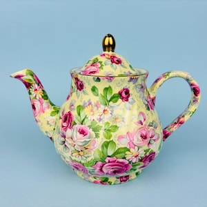 May include: A ceramic teapot with a pale yellow base colour featuring a repeating pattern of pink and white roses, green leaves, and small white flowers. The teapot has a curved handle, a spout, and a removable lid topped with a gold-coloured knob.