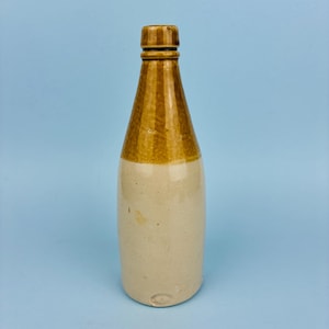 Peut inclure: Bouteille ancienne en grès avec un dégradé de couleurs beige et marron. La bouteille a une base arrondie et un col conique avec un dessus marron rayé. La partie inférieure est de couleur beige clair.