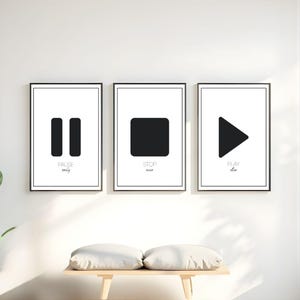 Op de afbeelding: Drie minimalistische kunstprints met zwarte symbolen op witte achtergronden. De prints tonen de pauze-, stop- en afspeelsymbolen, met bijbehorende tekst eronder. De lijsten zijn zwart en de prints worden boven een houten bank weergegeven.