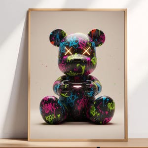 Op de afbeelding: Een ingelijste print met een zwarte teddybeerfiguur bedekt met kleurrijke graffiti-kunst. De beer heeft gloeiende gele kruisvormige ogen en houdt een gamecontroller vast. Het kunstwerk staat tegen een neutrale achtergrond.