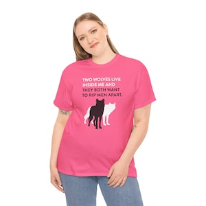 May include: A bright pink short-sleeve t-shirt featuring the white and black text: TWO WOLVES LIVE INSIDE ME AND THEY BOTH WANT TO RIP MEN APART. Below the text are two wolf silhouettes, one solid black and one solid white, standing side by side.