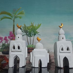 Può includere: Tre ornamenti in ceramica bianca a forma di moschea con lune crescenti dorate in cima. Gli ornamenti sono disposti in fila su una superficie di legno con uno sfondo tropicale.