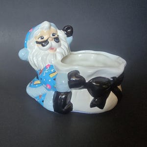 May include: A ceramic Santa Claus figurine in a seated position, holding a white sack with a black bow. Santa wears a blue suit with a white beard and black gloves. The planter is a festive holiday decoration.