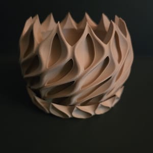 May include: A light brown, decorative, geometric vase with a unique, textured design. The vase features a series of repeating, curved, tear-drop shaped openings. The overall shape is round with a ridged top and bottom. The vase is made of a light brown material.