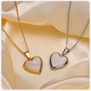 May include: Two heart-shaped pendant necklaces. One is gold-toned, the other silver-toned. Both have a white, pearlescent center. The pendants hang from delicate chains. The background is a soft, satin-like fabric.