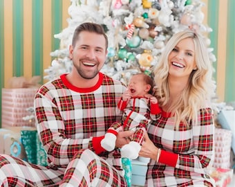 May include: A family wearing matching red, white, and green plaid pajamas poses in front of a decorated Christmas tree. The pajamas have red cuffs and trim. The family includes a baby, a man, and a woman.