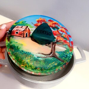 May include: A painted pot lid with a landscape scene featuring a red cabin, trees with red and orange leaves, and green foliage. A dark green handle with the word "imusa" is attached to the lid. The pot is silver.