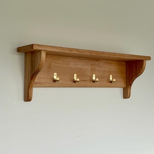 May include: Wooden wall shelf with four brass hooks. The shelf is made of light-colored wood and has a simple, functional design. The hooks are evenly spaced along the bottom edge, providing a place to hang items. The shelf is mounted on a white wall.