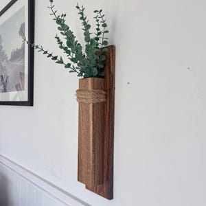 May include: A wall-mounted wooden vase featuring a rectangular dark brown wood block attached to a flat wooden backboard. The vase is accented with a thick, natural-coloured jute rope wrapped around the top and holds green eucalyptus stems.