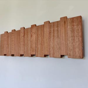 May include: A wall-mounted coat rack constructed from multiple vertical wooden slats of varying heights and widths. The natural light brown wood features a visible grain pattern, arranged in a staggered, overlapping design to create a textured surface.