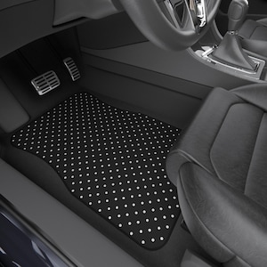 May include: Black car floor mat featuring a white polka dot design. The mat is rectangular with a curved edge, positioned in the driver's side footwell. The car interior is black, including a black leather seat.