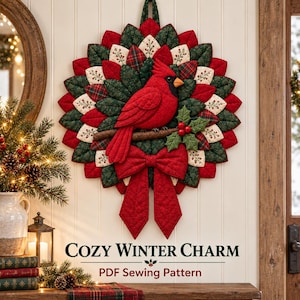 May include: A quilted fabric wall hanging featuring a red cardinal bird perched on a branch, surrounded by layered petals in red, dark green, and cream with holly berry accents. A large red fabric bow hangs at the bottom. Text reads COZY WINTER CHARM.