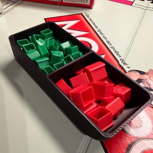 May include: A black plastic game piece organizer filled with green and red plastic houses. The organizer sits on a Monopoly game board. The game board has red and white text.