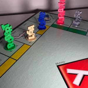 May include: A Monopoly board game with colorful player tokens spelling out names: Robert, Lisa, Edward, Elizabeth, and a silver token. The board features street names and a red corner with the word "BRAND".