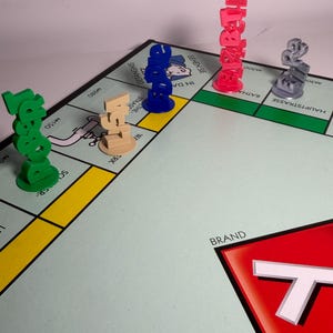 May include: A Monopoly board game with several colorful player tokens. The tokens are in green, tan, blue, red, and gray. The board has various property spaces and the Monopoly logo in the corner.