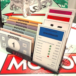Könnte beinhalten: Ein schwarzer Kartenhalter aus Kunststoff mit einem Stapel Monopoly-Besitzrechtkarten und Spielgeld. Die vordere Karte ist mit Schlossallee beschriftet und zeigt Mietwerte. Der Halter ist mit dem Namen Eddie in rotem Garn bestickt.
