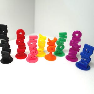 May include: A collection of colorful, 3D-printed name stands. Each stand features a different name in bold, stacked letters. Colors include black, red, pink, yellow, orange, green, purple, and blue. The stands are on a white surface.
