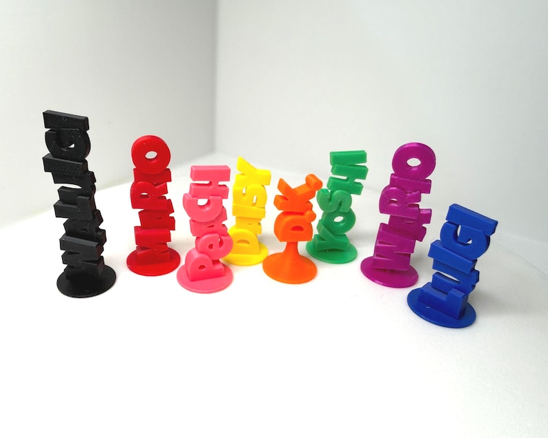 May include: A collection of colorful, 3D-printed name stands. Each stand features a different name in bold, stacked letters. Colors include black, red, pink, yellow, orange, green, purple, and blue. The stands are on a white surface.
