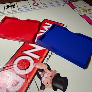 Könnte beinhalten: Zwei Kartenablagen aus Kunststoff, eine rote und eine blaue, liegen auf einem Monopoly-Spielbrett. Die Ablagen sind rechteckig mit einer gebogenen Vorderkante und einem Schlitz. Das Monopoly-Spielbrett ist im Hintergrund zu sehen.