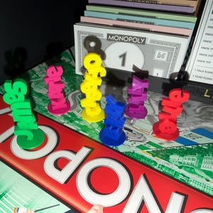 May include: A Monopoly board game with colorful player pieces spelling out names. The pieces are in green, pink, yellow, blue, purple, and red. A Monopoly money bill and property cards are also visible.
