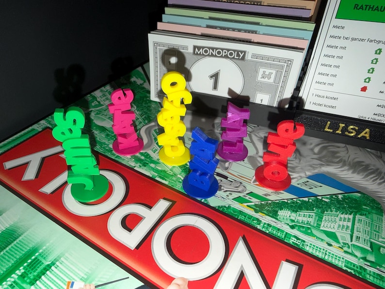 May include: A Monopoly board game with colorful player pieces spelling out names. The pieces are in green, pink, yellow, blue, purple, and red. A Monopoly money bill and property cards are also visible.