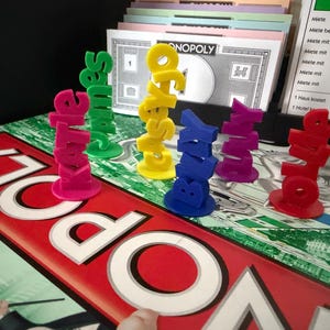 May include: A Monopoly board game with colorful, stacked name pieces spelling out names like "Kate" and "Sales". The game board is red and white, with the word "Monopoly" prominently displayed. Other game elements are visible.
