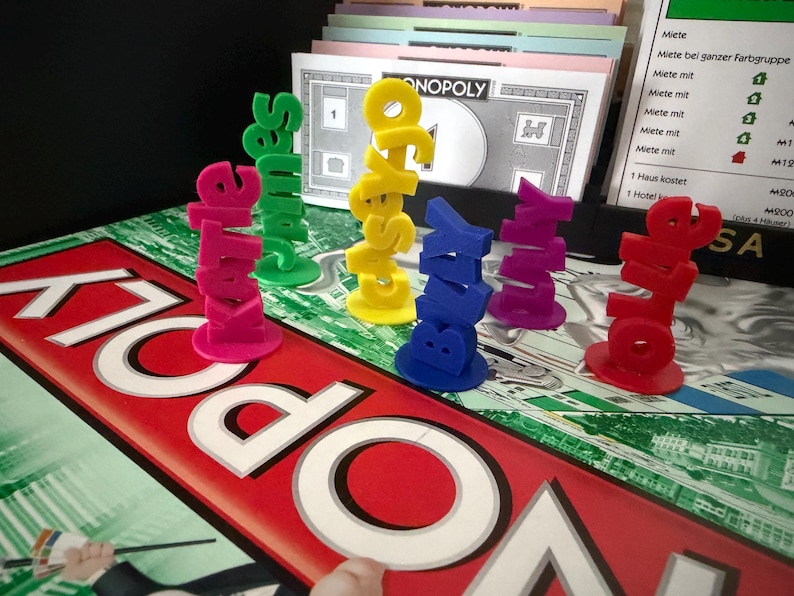 May include: A Monopoly board game with colorful, stacked name pieces spelling out names like "Kate" and "Sales". The game board is red and white, with the word "Monopoly" prominently displayed. Other game elements are visible.