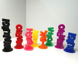 May include: A collection of colorful 3D-printed figurines, each spelling out a different name. The figurines are in various colors, including black, red, pink, yellow, orange, green, purple, and blue. Each figurine stands upright on a circular base.
