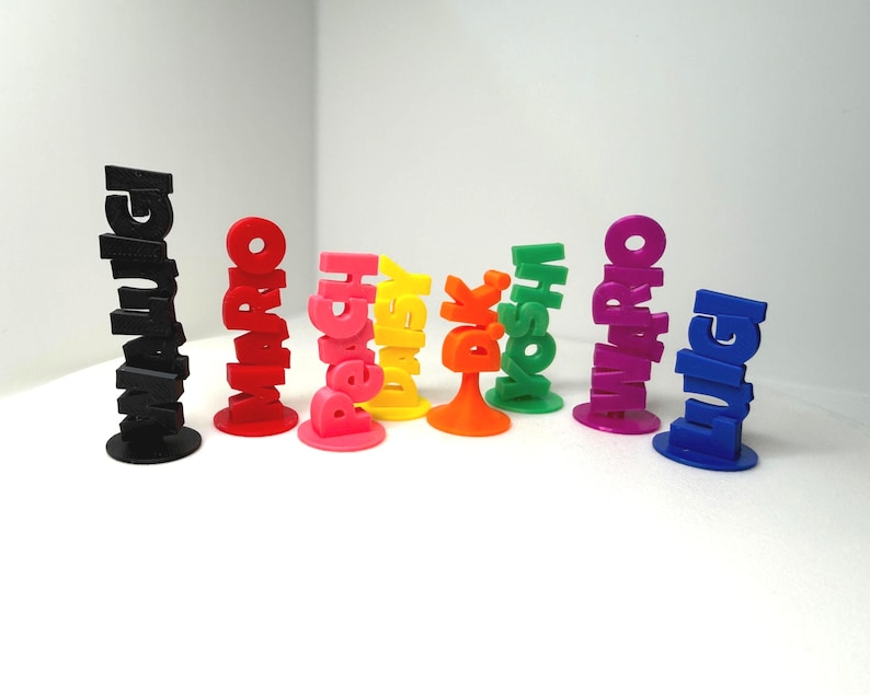 May include: A collection of colorful 3D-printed figurines, each spelling out a different name. The figurines are in various colors, including black, red, pink, yellow, orange, green, purple, and blue. Each figurine stands upright on a circular base.