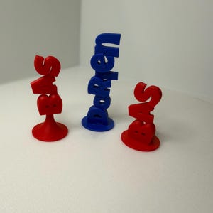 May include: Three 3D-printed objects. Two are red, shaped like the letters "S" and "B" stacked, with a base. The third is blue, with the word "Doreen" printed vertically. All objects are on a white surface.