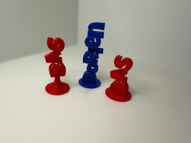 May include: Three 3D-printed objects. Two are red, shaped like the letters "S" and "B" stacked, with a base. The third is blue, with the word "Doreen" printed vertically. All objects are on a white surface.