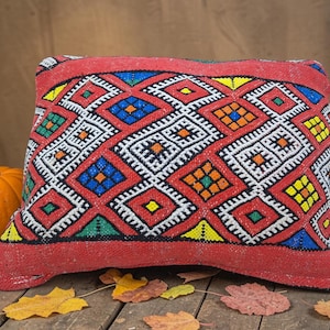 May include: A rectangular red pillow with a geometric pattern in white, blue, green, orange, and yellow. The pillow is placed on a wooden surface with a small pumpkin and autumn leaves.