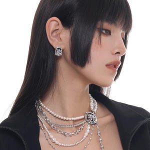 Puede incluir: Un collar de varias hebras con perlas de imitación, cadenas plateadas y un colgante decorativo. El collar se combina con pendientes a juego. La joyería se lleva sobre una chaqueta negra.