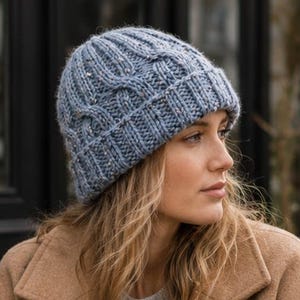 May include: A light blue cable-knit beanie with a ribbed cuff and subtle flecked texture, worn with a tan wool-blend coat, a grey sweater, and blue denim jeans, while holding a disposable paper coffee cup with a white plastic lid.