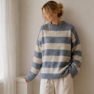 May include: A blue and cream striped sweater with a crew neck. The sweater has wide horizontal stripes and long sleeves. The model is wearing white trousers. The sweater is made of a soft, textured material.