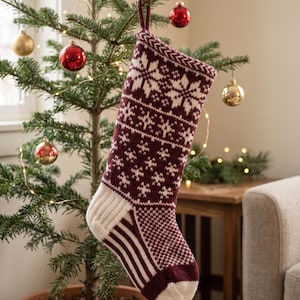 May include: A knitted Christmas stocking featuring a deep burgundy and cream color palette. The design includes intricate snowflake patterns across the upper section, with a ribbed cream heel and toe, and vertical stripes along the foot area.
