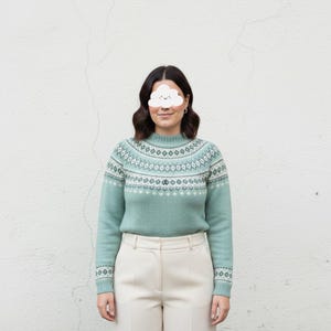 May include: A light teal sweater with a high neckline and a detailed geometric pattern in white and light blue. The sweater is paired with cream-colored pants. The overall style is casual and comfortable, suitable for cooler weather.