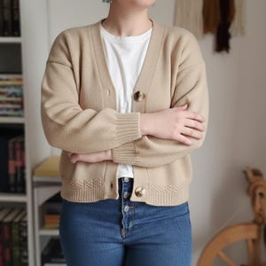 以下が含まれることがあります： Vネックとボタン留めのベージュカーディガン。長袖セーターは白いTシャツとブルージーンズと合わせています。カーディガンはリブ編みの袖口と、裾に装飾的なニット模様が施されています。