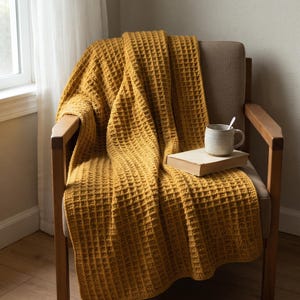 May include: A mustard yellow waffle-knit throw blanket draped over a wooden armchair. A closed hardcover book and a ceramic mug with a spoon inside are placed on top of the blanket. The blanket features a textured grid pattern throughout.