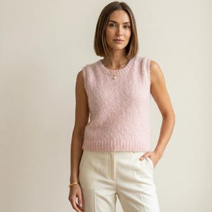 May include: A pale pink sleeveless sweater vest with a textured finish. The vest has a crew neck and is worn with white trousers. The model is wearing a gold bracelet. The background is a plain, neutral colour.