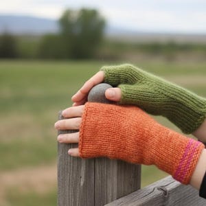 Puede incluir: Guantes sin dedos de punto en tonos verde y naranja. El guante naranja tiene un puño a rayas rosas y amarillas. Los guantes descansan sobre un poste de valla de madera, con un campo verde borroso y montañas al fondo.