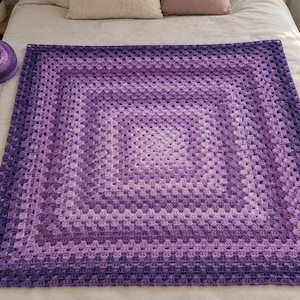 Könnte beinhalten: Eine quadratische Häkeldecke mit Granny-Square-Muster in einem Farbverlauf aus verschiedenen Violetttönen, der von hellem Lavendel in der Mitte bis zu tiefem Violett an den Rändern reicht. Ein passendes Knäuel violettes Garn liegt daneben.