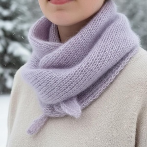 Può includere: Sciarpa lilla lavorata a maglia con un motivo strutturato. La sciarpa è avvolta attorno al collo e presenta un piccolo dettaglio a fiocco. Lo sfondo mostra una scena esterna innevata, adatta all'inverno.