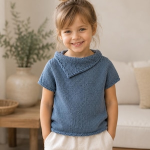 May include: A short-sleeved, blue knitted pullover featuring a distinctive folded cowl neckline. The garment is crafted from a textured yarn with a visible stockinette stitch pattern, finished with a simple ribbed hem and sleeve edges.