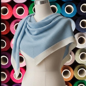 Könnte beinhalten: Ein hellblaues Tuch mit weißem Rand ist auf einer Schaufensterpuppe drapiert. Der Hintergrund ist eine Wand aus bunten Garnrollen in Rosa, Grün, Blau und Lila. Das Tuch ist aus weichem Strickstoff.