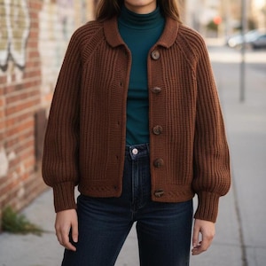Könnte beinhalten: Ein brauner Cardigan mit Knöpfen, Kragen und Rippenstruktur. Der langärmlige Pullover hat eine lockere Passform und wird mit einem türkisfarbenen Rollkragenpullover und dunkelblauen Jeans kombiniert. Die Knöpfe sind farblich passend braun.