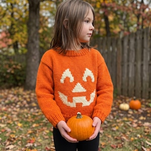 Puede incluir: Un suéter de punto de manga larga en color naranja brillante con un gráfico de cara de calabaza de Halloween en color crema en la parte delantera. El suéter tiene un cuello redondo acanalado y puños acanalados, con textura de punto visible.