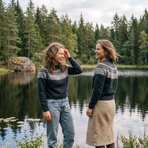 May include: Two people wearing matching dark blue sweaters with a white and gray geometric pattern across the chest and shoulders. One person wears blue jeans, the other a tan skirt. They stand near a lake with a forest backdrop.